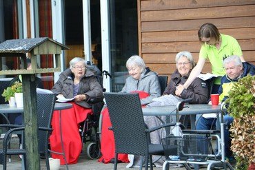 Ældre der sidder udenfor med tæpper over sig og får serveret kaffe af personale